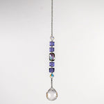 Small sun catcher hanging vertically against blurred background, made with purple crystal cube beads, a large emerald cut crystal and a round crystal prism anchor.