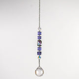 Small sun catcher hanging vertically against blurred background, made with purple crystal cube beads, a large emerald cut crystal and a round crystal prism anchor.