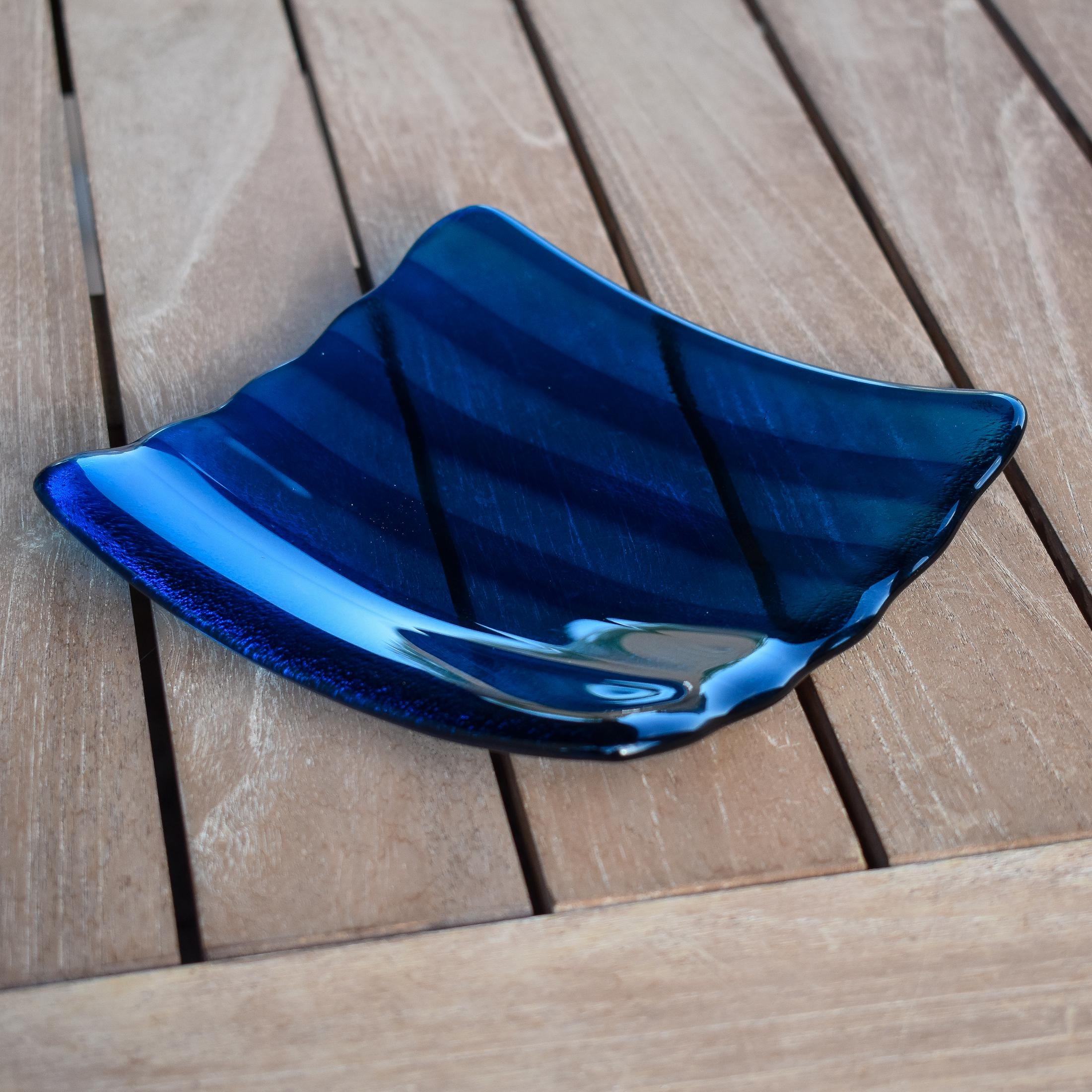 Deep aqua blue fused glass tray with two scalloped edges and strips of a lighter color. The tray is sitting on a wood table at an angle, outdoors, with some reflections in the glass.