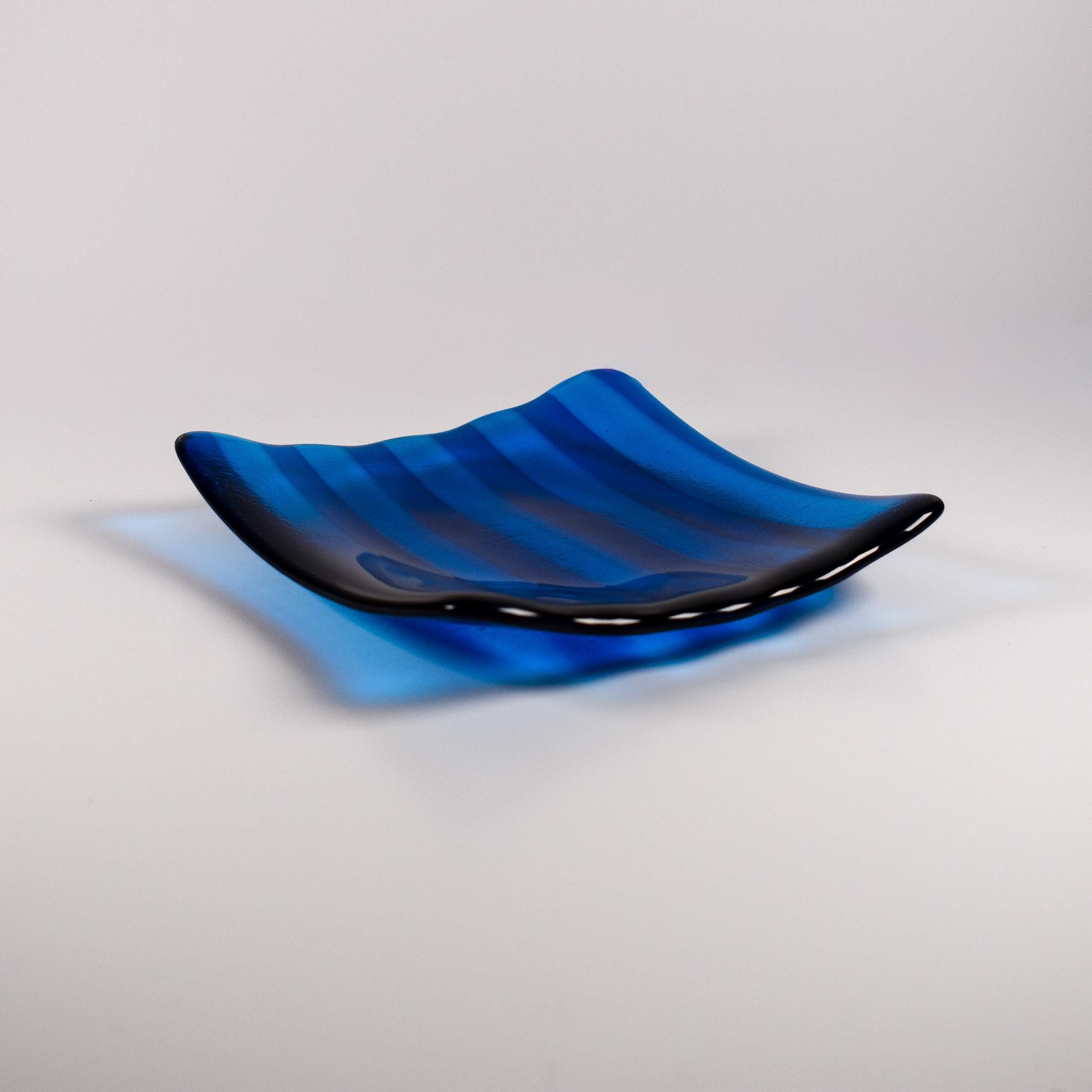 Deep aqua blue fused glass tray with two opposite scalloped edges and strips of a lighter color. The tray is sitting at an angle on a white surface.