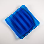 Deep aqua blue fused glass tray with two opposite scalloped edges and strips of a lighter color. The photo is looking down from the top of the tray. The tray is sitting on a white surface.