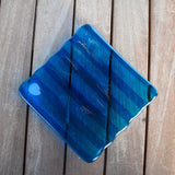 Deep aqua blue fused glass tray with two opposite scalloped edges and strips of a lighter color. The photo is looking down from the top of the tray. The tray is sitting on a wood table. Photo was taken outdoors and there is some sunlight reflection on the glass.