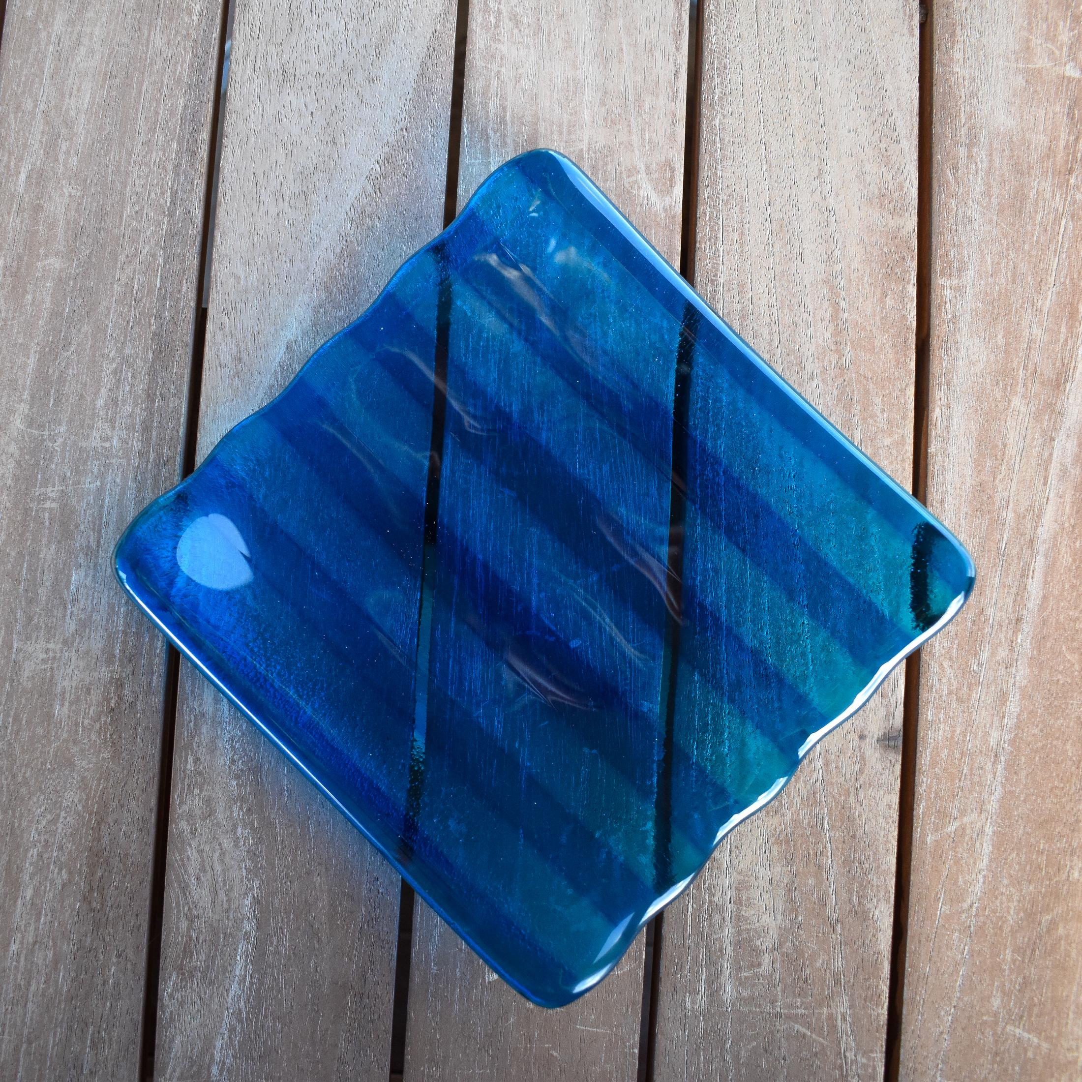Deep aqua blue fused glass tray with two opposite scalloped edges and strips of a lighter color. The photo is looking down from the top of the tray. The tray is sitting on a wood table. Photo was taken outdoors and there is some sunlight reflection on the glass.