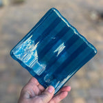 Deep aqua blue fused glass tray with two opposite scalloped edges and strips of a lighter color being hand-held up into sunlight. Photo was taken outdoors and there is some sunlight reflection on the glass.