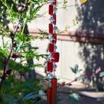 Two ends of a long stainless steel wire, hanging vertically with handmade fused glass rectangular beads in red, paired with clear glass beads. Each end of the wire is anchored by a long rectangular piece of red fused glass. Hanging in a garden.