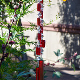 Two ends of a long stainless steel wire, hanging vertically with handmade fused glass rectangular beads in red, paired with clear glass beads. Each end of the wire is anchored by a long rectangular piece of red fused glass. Hanging in a garden.