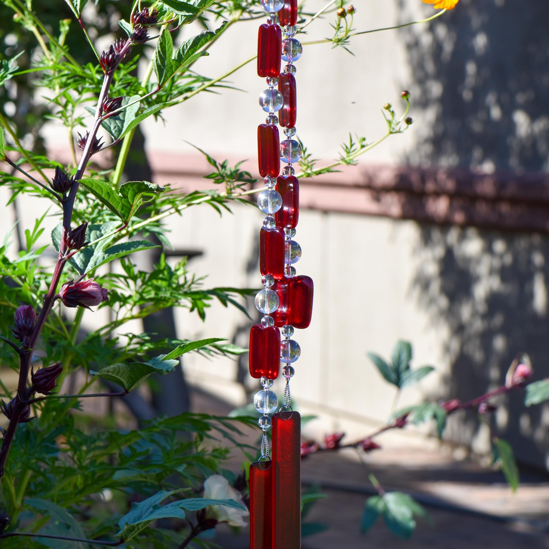 Two ends of a long stainless steel wire, hanging vertically with handmade fused glass rectangular beads in red, paired with clear glass beads. Each end of the wire is anchored by a long rectangular piece of red fused glass. Hanging in a garden.
