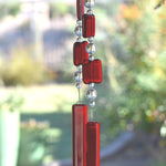Two ends of a long stainless steel wire, hanging vertically with handmade fused glass rectangular beads in red, paired with clear glass beads. Each end of the wire is anchored by a long rectangular piece of red fused glass. Blurred foliage in background.