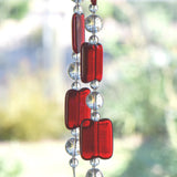 Two ends of a long stainless steel wire, hanging vertically with handmade fused glass rectangular beads in red, paired with clear glass beads. Blurred foliage in background.