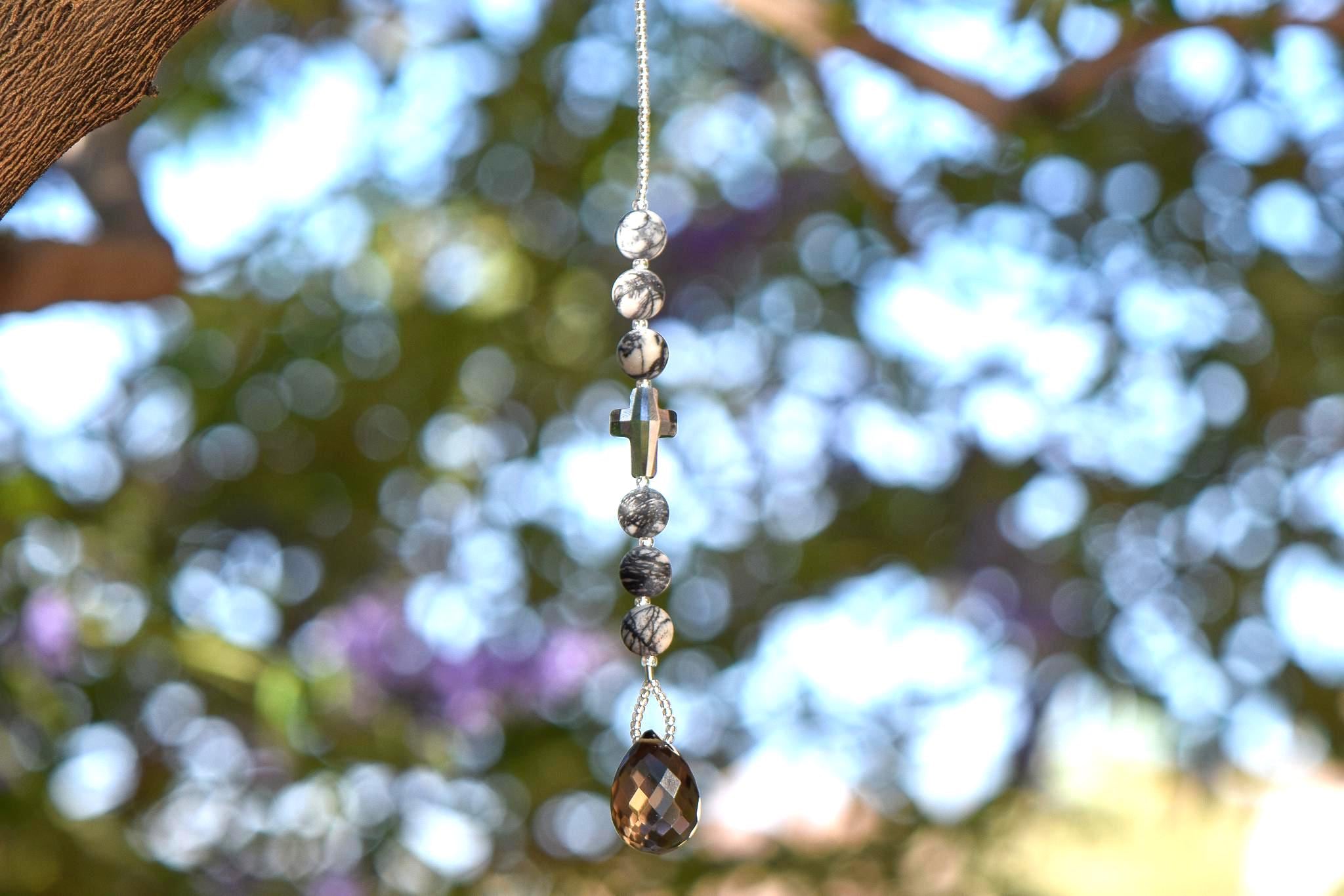 Small sun catcher hanging vertically against blurred foliage background. Silver-gray crystal cross is flanked by six jasper stone beads in a marbled gray, anchored by large smoky quartz pendant