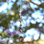 Small sun catcher hanging vertically against blurred foliage background. Silver-gray crystal cross is flanked by six jasper stone beads in a marbled gray, anchored by large smoky quartz pendant