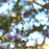 Small sun catcher hanging vertically against blurred foliage background. Silver-gray crystal cross is flanked by six jasper stone beads in a marbled gray, anchored by large smoky quartz pendant