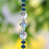 Close up view of crystal cross bead strung on wire, hanging vertically. The cross bead is flanked by two round, faceted glass crystal beads and small rondelle turquoise-colored beads.