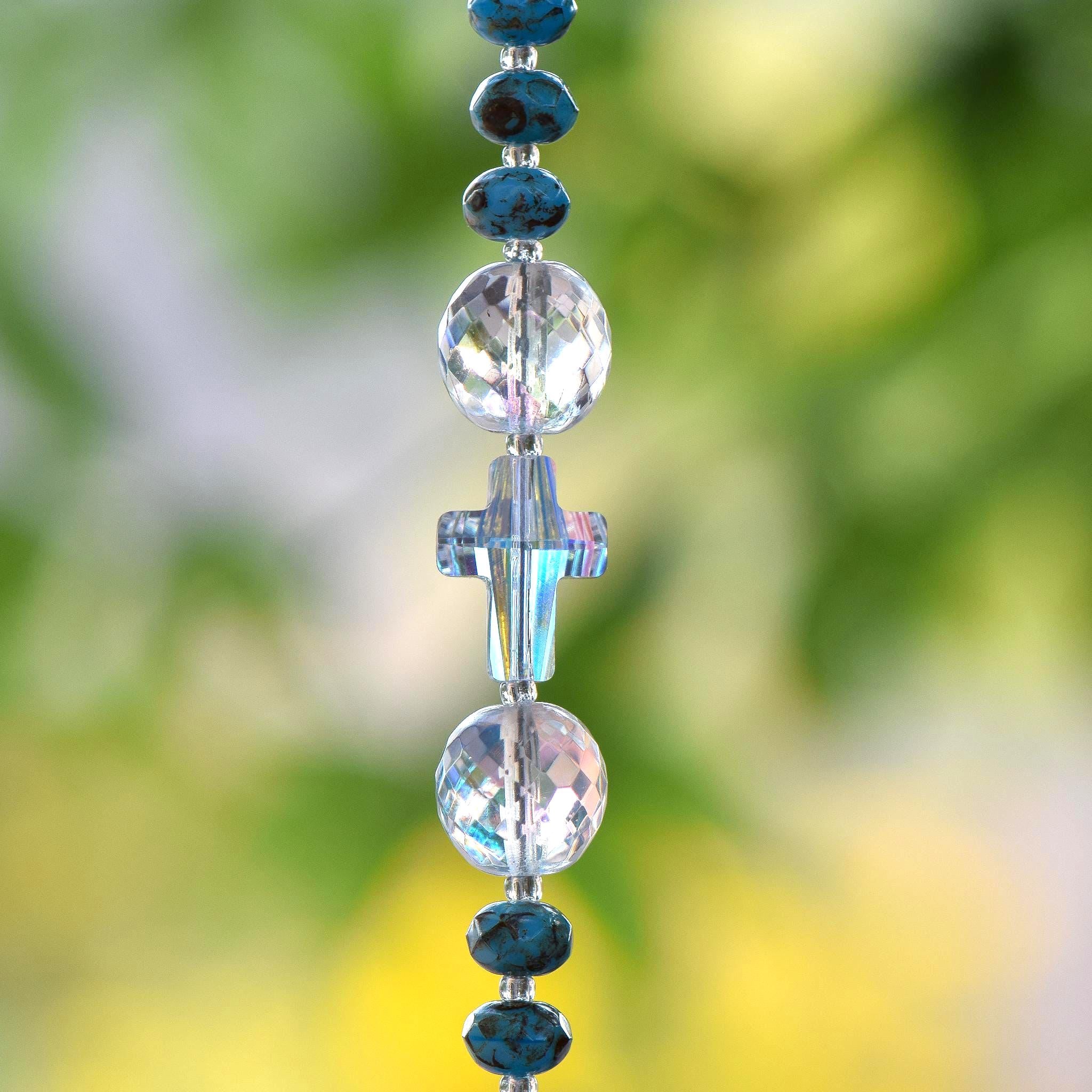 Close up view of crystal cross bead strung on wire, hanging vertically. The cross bead is flanked by two round, faceted glass crystal beads and small rondelle turquoise-colored beads.