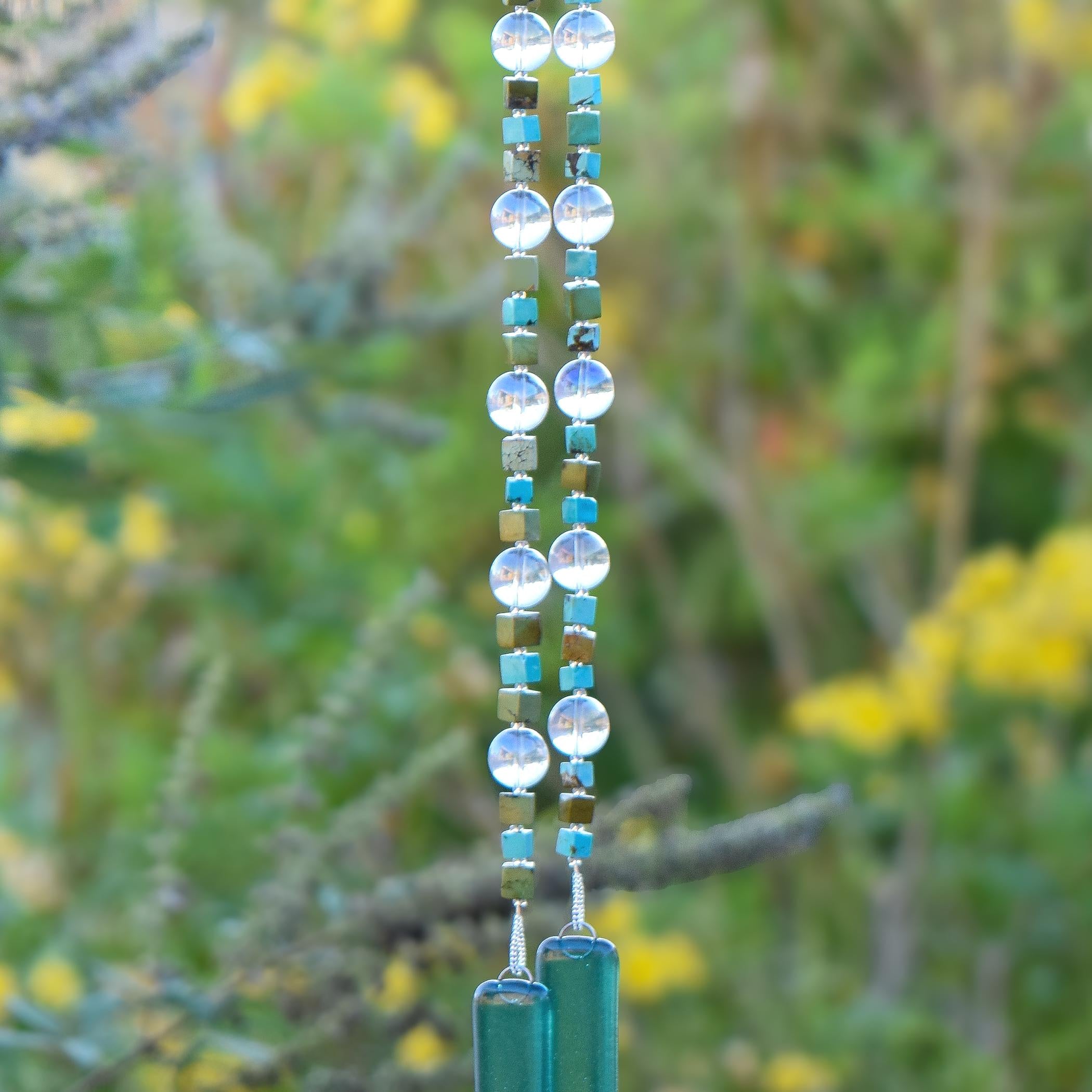 Two ends of a long wire hanging down covered in beads with glass anchors. The beads include cubic stones of howlite or magnesite, large clear glass beads with a reflective finish and glass silver-lined seed beads. The anchor glass is kiln formed, caribbean blue transparent. Background is blurred foliage.