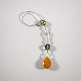 Glass crystal cross is beaded with other crystal beads and anchored by Carnelian Agate stone pendant to make a small sun catcher for hanging in a window or from the rear view mirror inside a car. The sun catcher is laying flat in a white surface.