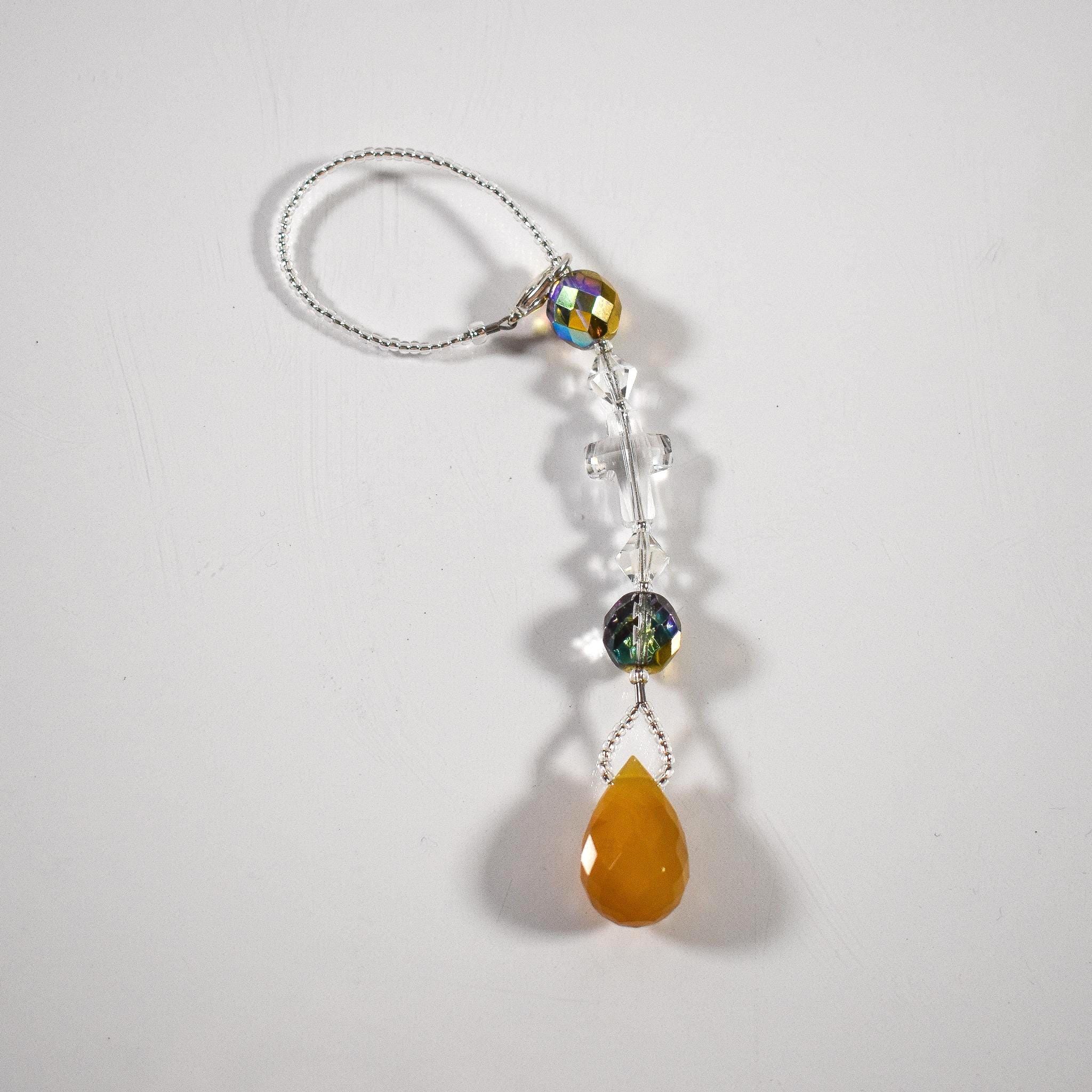 Glass crystal cross is beaded with other crystal beads and anchored by Carnelian Agate stone pendant to make a small sun catcher for hanging in a window or from the rear view mirror inside a car. The sun catcher is laying flat in a white surface.