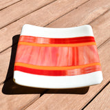 Square fused glass tray with vanilla cream colored base and a striped and streaky design of red, orange and white glass. Tray is 6&quot; square with upturned corners. Sitting on a wood table in the sunlight. View is from the top and the side.