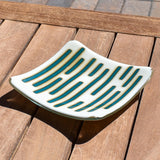 Square glass dish, viewed from above and front, on a slatted wood table. The dish has a base color of vanilla cream with abstract design consisting of lines of peacock green.