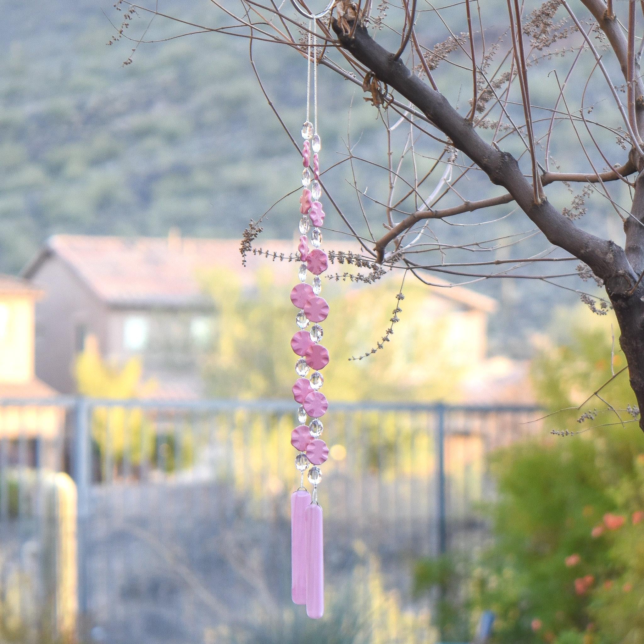 Pink magnesite stone beads shaped as wavy flat rounds are strung with glass crystal ovals and anchored by two pieces of pink fused glass. Hanging from tree. Photo shows the entire chime from where the wire is hung from a hook to the bottom of the two glass pieces. Background is a home in the distance in front of a mountain.