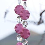 Four pink magnesite stone beads shaped as wavy, flat rounds, are two strings of wire with oval glass crystal beads. The strings are hanging in front of a mountain in the background.
