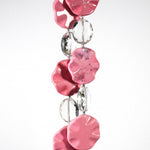 Six pink magnesite stone beads shaped as wavy, flat rounds, on two strings of wire with oval glass crystal beads. Hanging in front of a white surface.