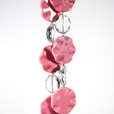 Six pink magnesite stone beads shaped as wavy, flat rounds, on two strings of wire with oval glass crystal beads. Hanging in front of a white surface.