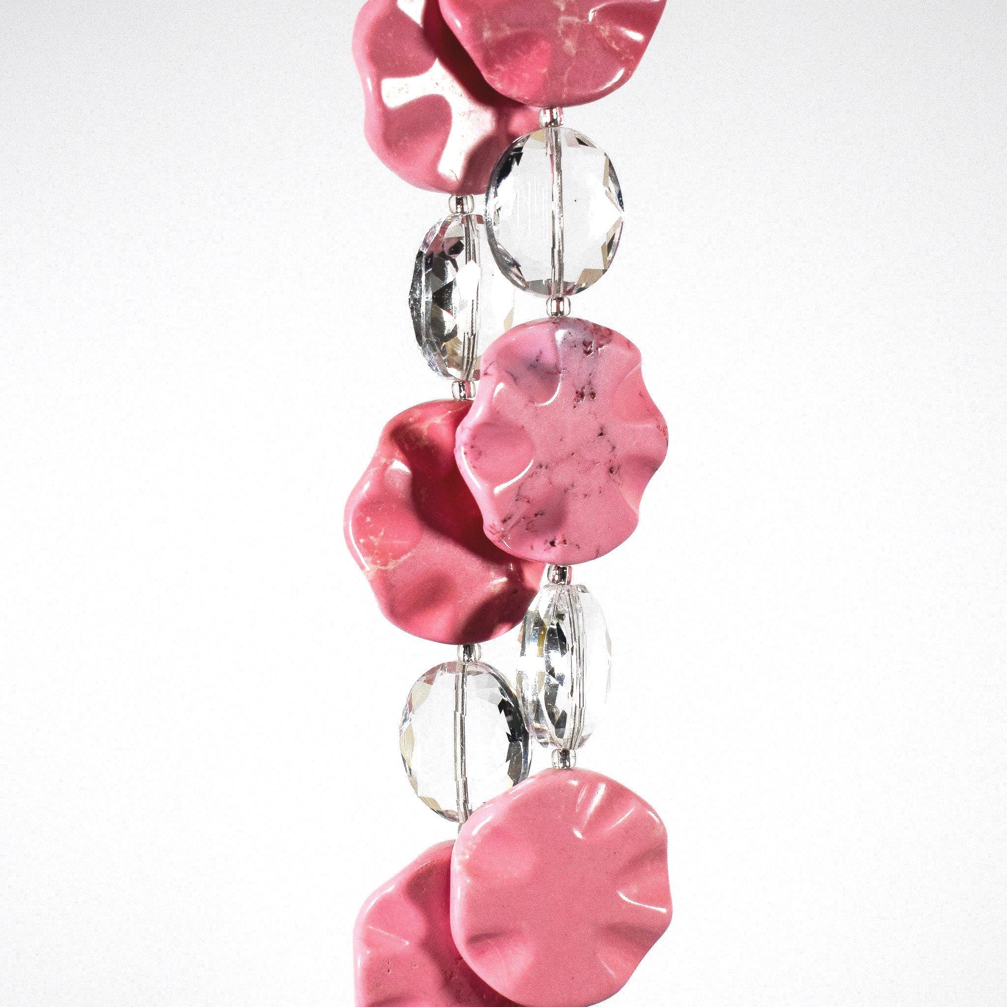 Six pink magnesite stone beads shaped as wavy, flat rounds, on two strings of wire with oval glass crystal beads. Hanging in front of a white surface.