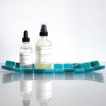 A rectangular glass tray with a mosaic tile design. The tray features a pattern of square tiles in shades of teal and white. The tiles are arranged in a grid-like pattern, creating a visually appealing surface. The tray is placed on a white surface. Personal care bottles are sitting on top of the tray.
