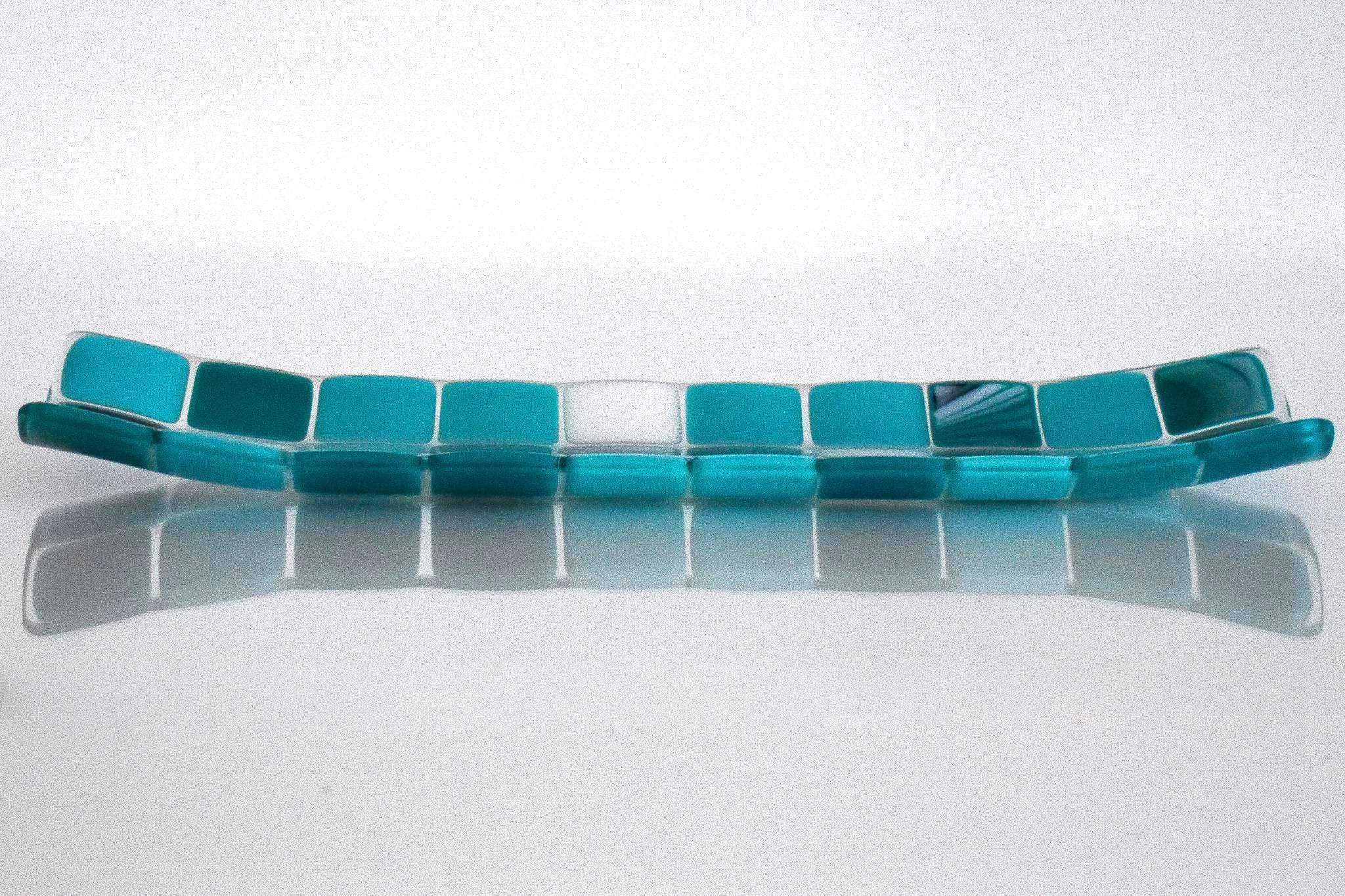 A rectangular glass tray with a mosaic tile design. The tray features a pattern of square tiles in shades of teal and white. The tiles are arranged in a grid-like pattern, creating a visually appealing surface. The tray is placed on a white surface. View is from the long side.