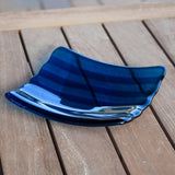 Deep aqua blue fused glass tray with two scalloped edges and strips of a lighter color. The tray is sitting on a wood table at an angle, outdoors, with some reflections in the glass.
