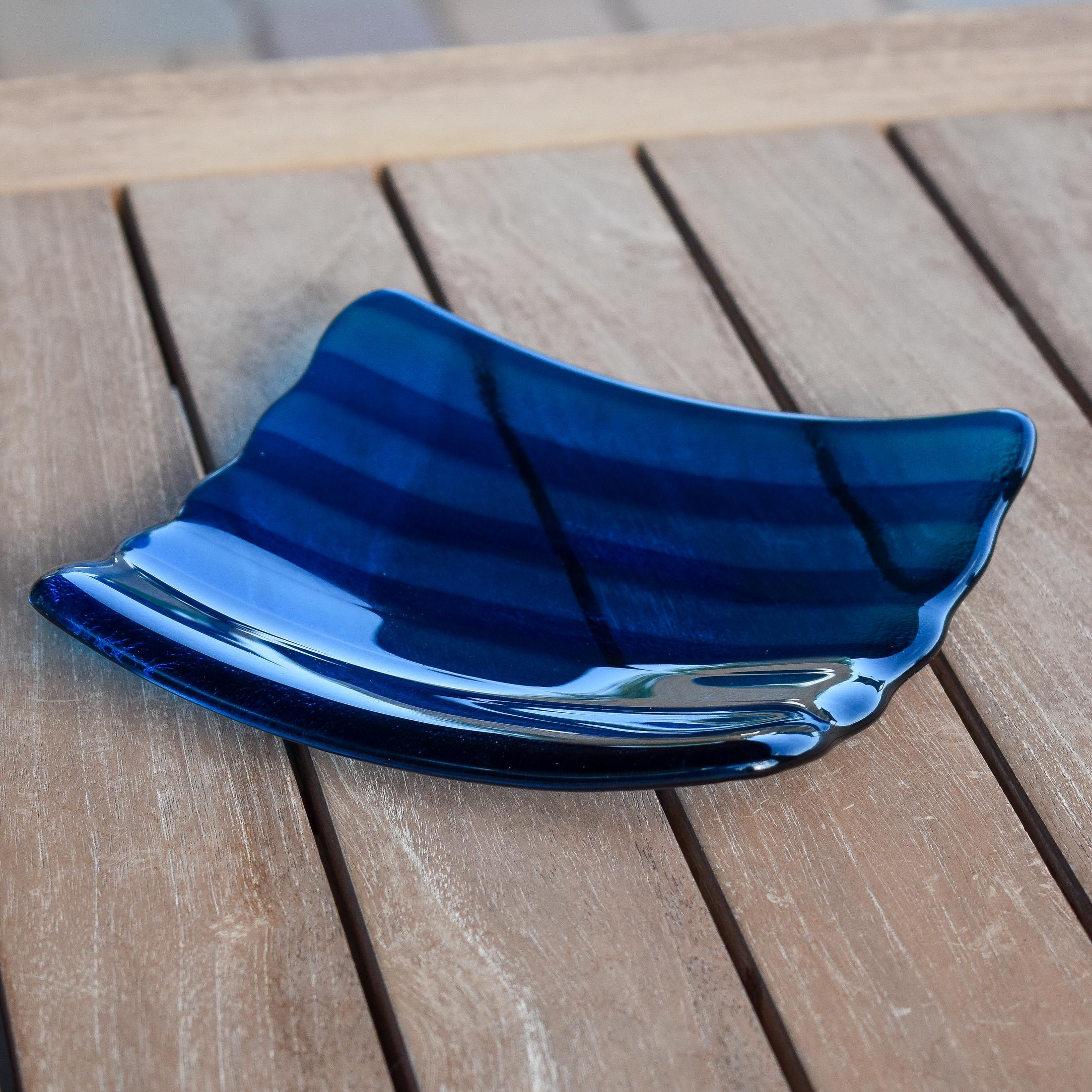 Deep aqua blue fused glass tray with two scalloped edges and strips of a lighter color. The tray is sitting on a wood table at an angle, outdoors, with some reflections in the glass.
