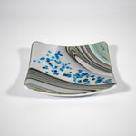 Six inch square glass tray with modern design on wood table.Tray is white with swirls and flakes of aqua, cream, tan and gray. On a white surface.