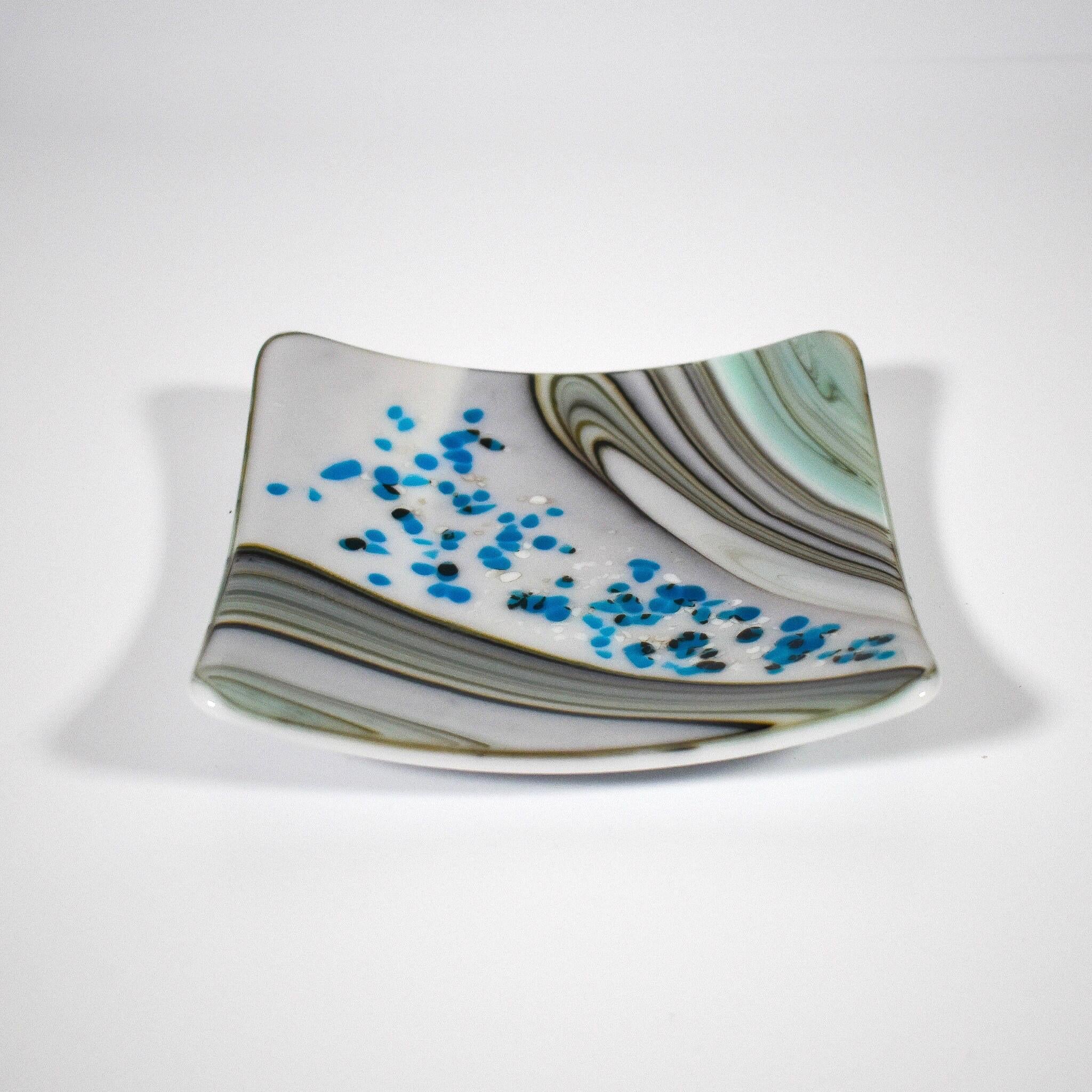 Six inch square glass tray with modern design on wood table.Tray is white with swirls and flakes of aqua, cream, tan and gray. On a white surface.