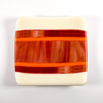 Square fused glass tray with vanilla cream colored base and a striped and streaky design of red, orange and white glass. Tray is 6&quot; square with upturned corners. Camera is looking straight down above the tray which is sitting on a white surface.