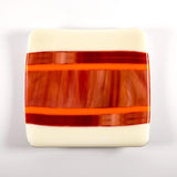 Square fused glass tray with vanilla cream colored base and a striped and streaky design of red, orange and white glass. Tray is 6&quot; square with upturned corners. Camera is looking straight down above the tray which is sitting on a white surface.