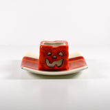 Square fused glass tray with vanilla cream colored base and a striped and streaky design of red, orange and white glass. A small pumpkin candle holder is sitting in the tray to show how it can be used. Tray is 6&quot; square with upturned corners. Sitting on a white surface.