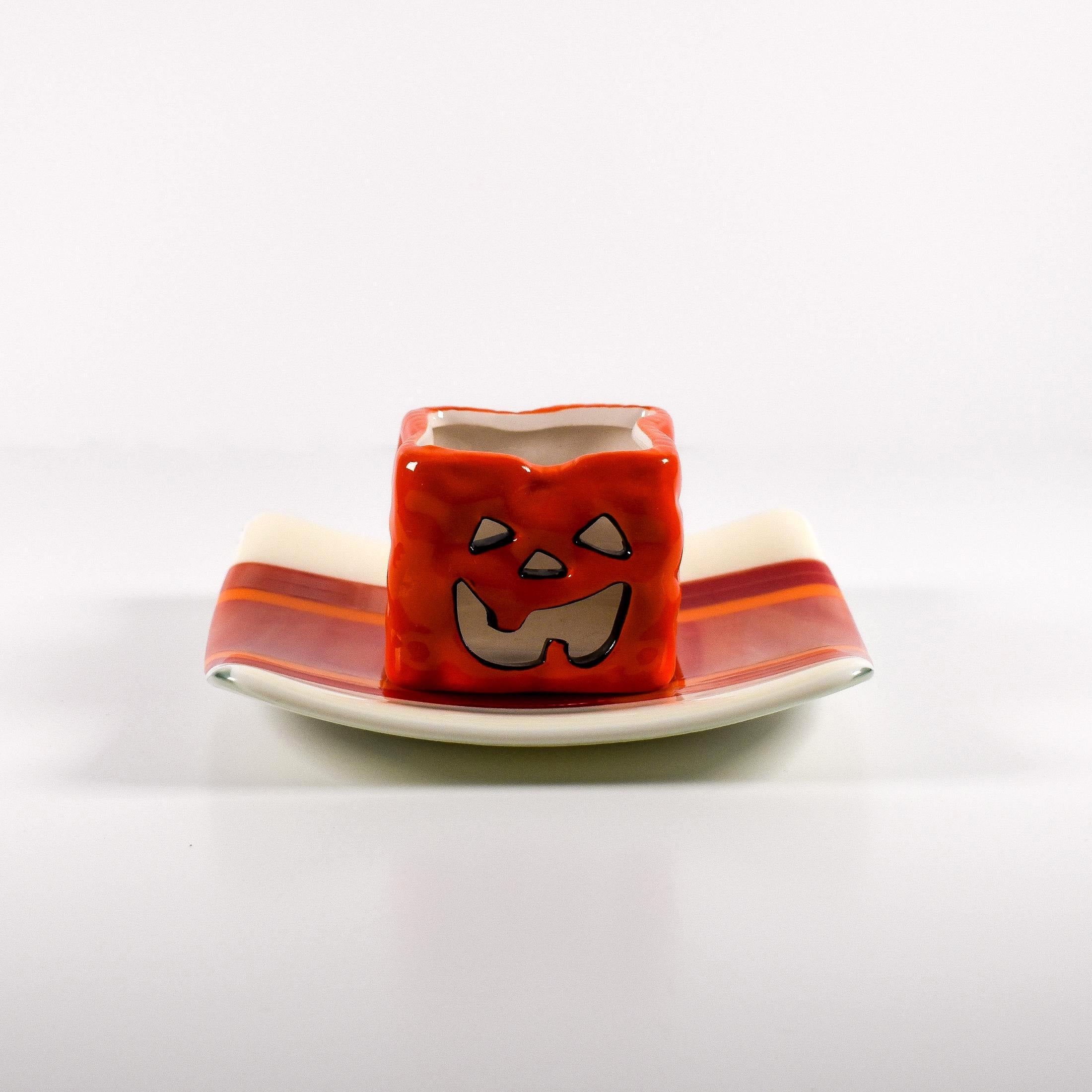 Square fused glass tray with vanilla cream colored base and a striped and streaky design of red, orange and white glass. A small pumpkin candle holder is sitting in the tray to show how it can be used. Tray is 6&quot; square with upturned corners. Sitting on a white surface.