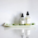 Channel-shaped rectangular fused glass tray in vanilla cream with green and yellow accents that look like lime slices. Viewed from the long side. Sitting on a white reflective surface. Tray contains two skin care products in clear bottles.