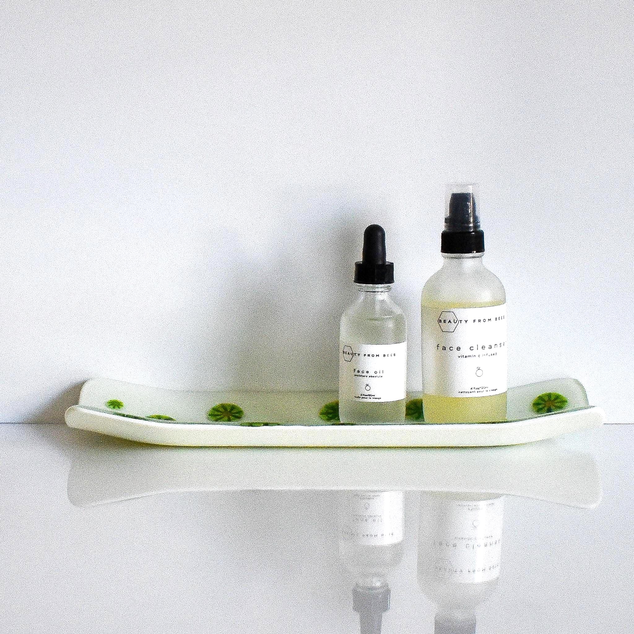Channel-shaped rectangular fused glass tray in vanilla cream with green and yellow accents that look like lime slices. Viewed from the long side. Sitting on a white reflective surface. Tray contains two skin care products in clear bottles.