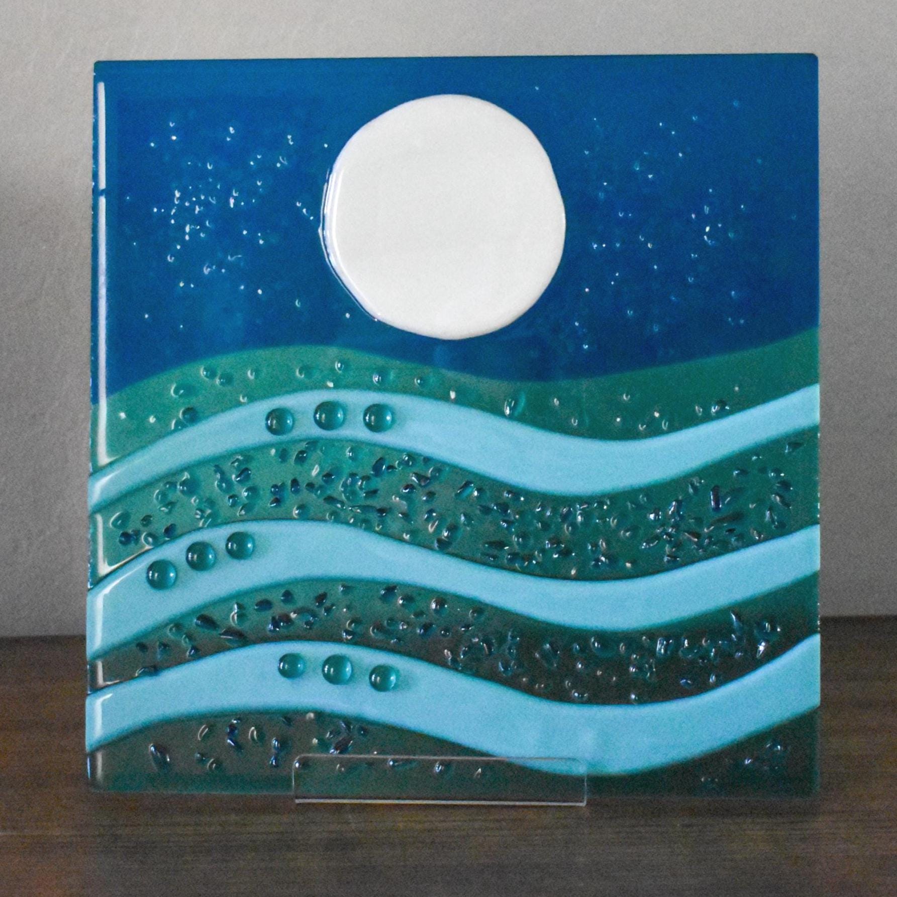 A 10 inch by 10 inch square glass panel on a clear acrylic stand sitting on a wood table in front of a light beige wall. The panel is depicting  a moon and stars against an aqua blue piece of glass. At the bottom are wave patterns made with caribbean blue glass, turquoise blue glass and seafoam green glass dots.