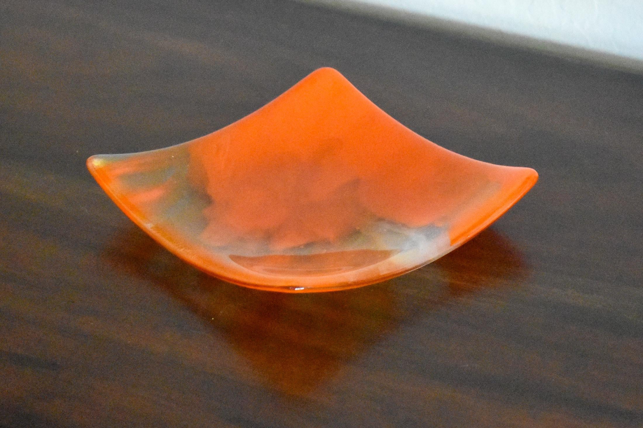 Square fused glass tray made of clear glass with streaks of bright orange, viewed from an agle as it is sitting on a dark brown wood table.