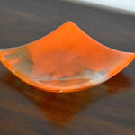 Square fused glass tray made of clear glass with streaks of bright orange, viewed from an agle as it is sitting on a dark brown wood table.