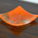 Square fused glass tray made of clear glass with streaks of bright orange, viewed from an agle as it is sitting on a dark brown wood table.