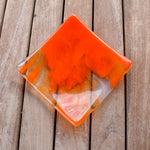 Square fused glass tray made of clear glass with streaks of bright orange, viewed from above as it is sitting on a slatted wood table.