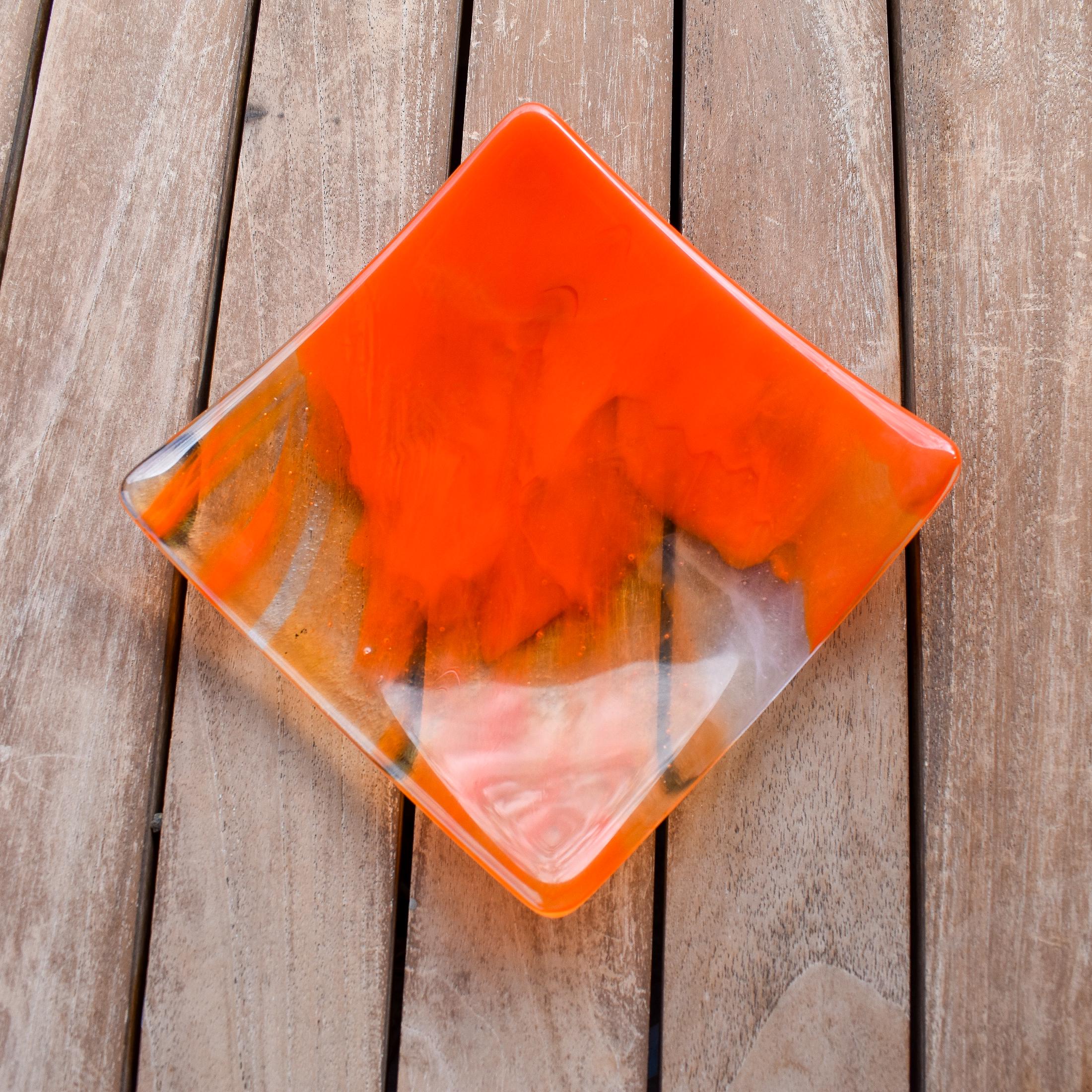 Square fused glass tray made of clear glass with streaks of bright orange, viewed from above as it is sitting on a slatted wood table.