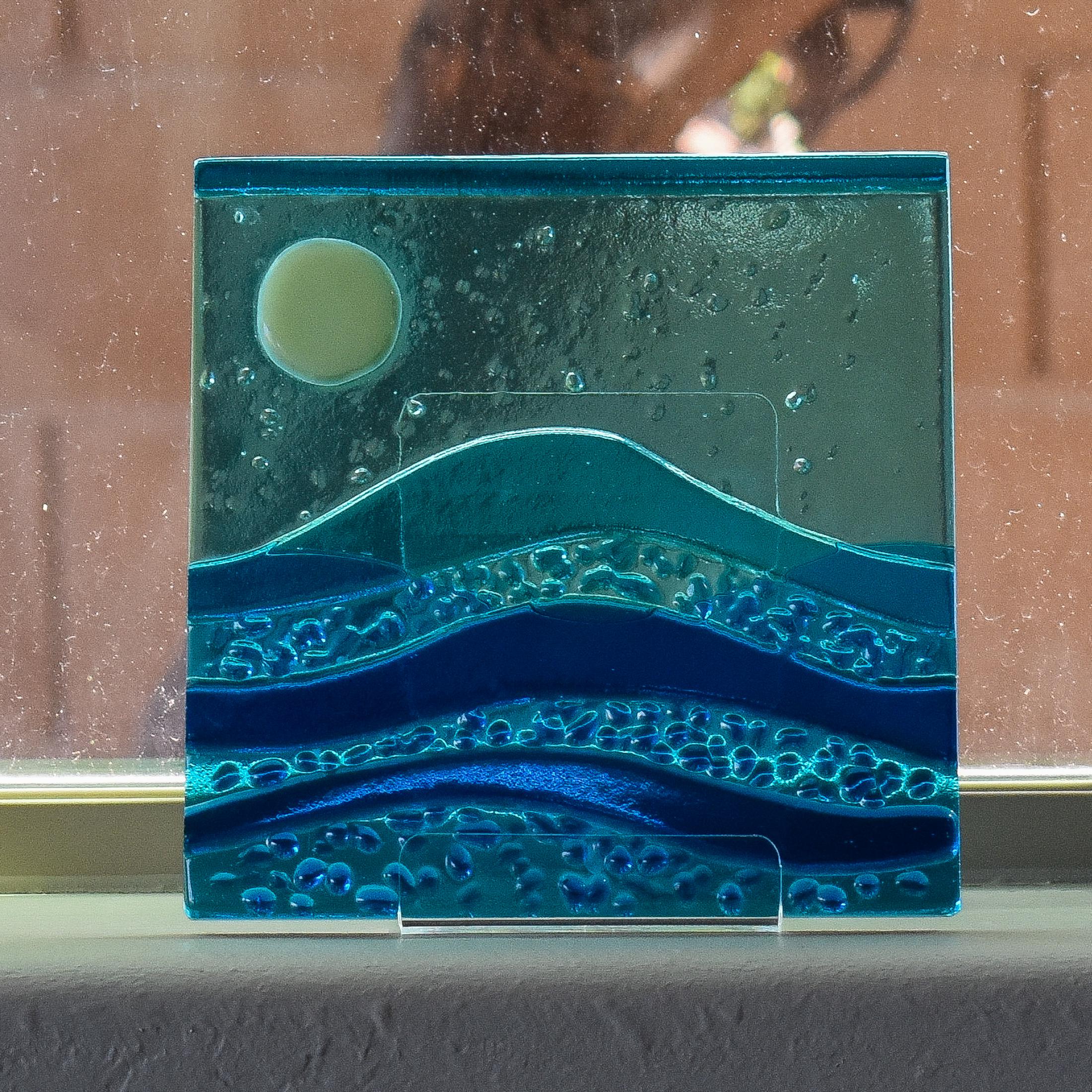 Six inch square glass panel on a window sill with a tree trunk behind. The panel depicts waves of blue with texture and flow underneath a full moon in an evening sky.