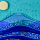 Close up of six inch square glass panel on a clear acrylic stand on a white background. The panel depicts waves of blue with texture and flow underneath a full moon in an evening sky.