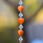 MA string of orange heart-shaped beads with clear crystal beads in between. The beads are strung on a stainless steel wire and hanging vertically in front of blurred background.
