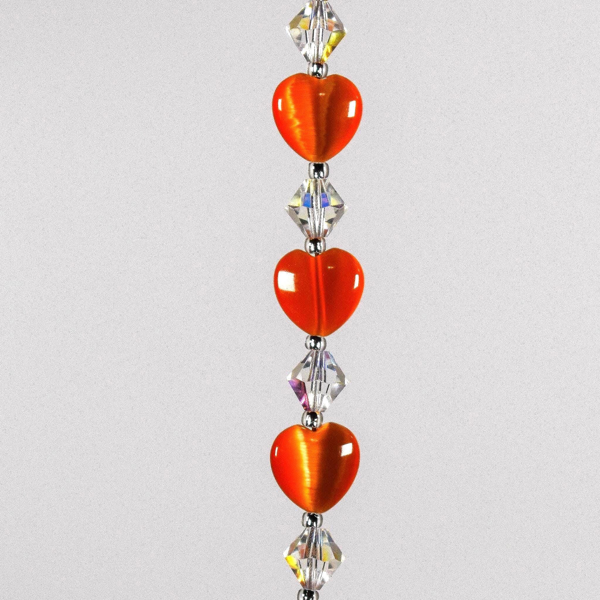 Three orange heart-shaped glass beads with small glass crystal beads in between, strung on stainless steel, hanging vertically against white background.